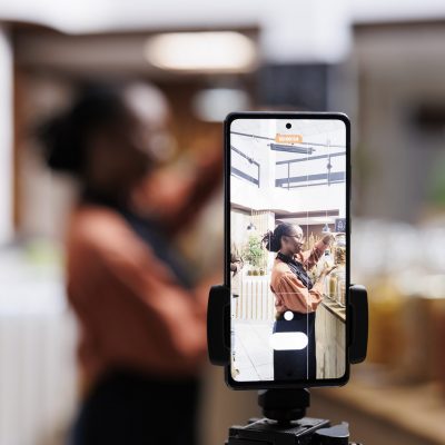 The eco-conscious owner promotes zero waste with reusable packaging. Social media vlogger films using a smartphone and tripod. Vibrant shelves and a camera capture the moment. Focus on the foreground.